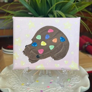 May include: A small, square canvas painting featuring a brown, abstract shape with colorful gem-like accents. The background is light pink with yellow stars. The artwork is displayed on a clear easel, set on a white, decorative plate.