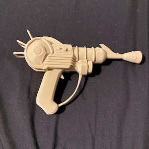 May include: A beige ray gun with a detailed design, featuring a circular front, a ribbed body, and a handle. The toy gun has a trigger and a curved wire detail. The overall design suggests a retro or science fiction theme.