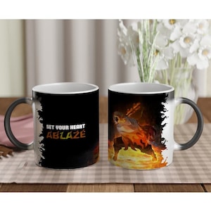 Op de afbeelding: Een zwarte keramische mok met een afbeelding van een personage omringd door oranje en gele vlammen. De tekst "SET YOUR HEART ABLAZE" staat in het wit en oranje op de zijkant gedrukt. De mok heeft een zwart handvat en een witte binnenrand.