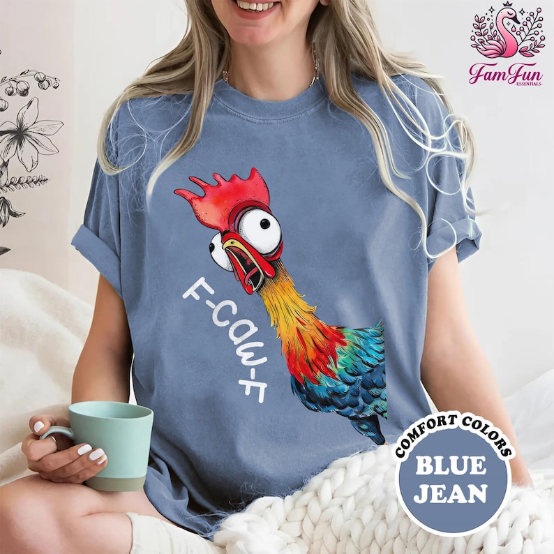 A woman wearing a blue t-shirt with a colorful cartoon rooster graphic and the text "F-CauT" printed on it. She is sitting on a bed and holding a mug.