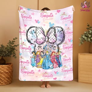 A person is holding up a pink and white blanket with a Disney princess theme, featuring illustrations of various Disney princesses.