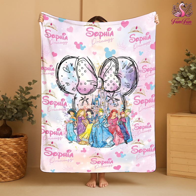 A person is holding up a pink and white blanket with a Disney princess theme, featuring illustrations of various Disney princesses.