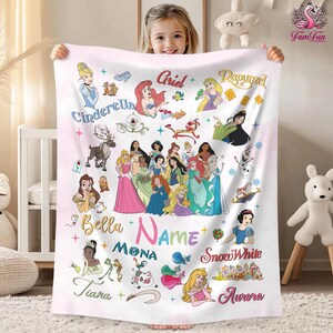 A young girl is holding a blanket featuring various Disney princess characters, including Cinderella, Belle, Mona, Snow White, and Aurora.
