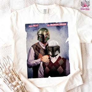 A white t-shirt with a printed image of two men wearing Star Wars-style helmets, one in a green helmet and the other in a silver helmet, standing together.