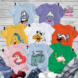 May include: A collection of eight short-sleeved t-shirts in various colors, each featuring a cartoon character from a popular animated film. The shirts are light blue, purple, yellow, white, orange, pink, light blue, and green. The characters include a smiling fish, a sea witch, and a king.