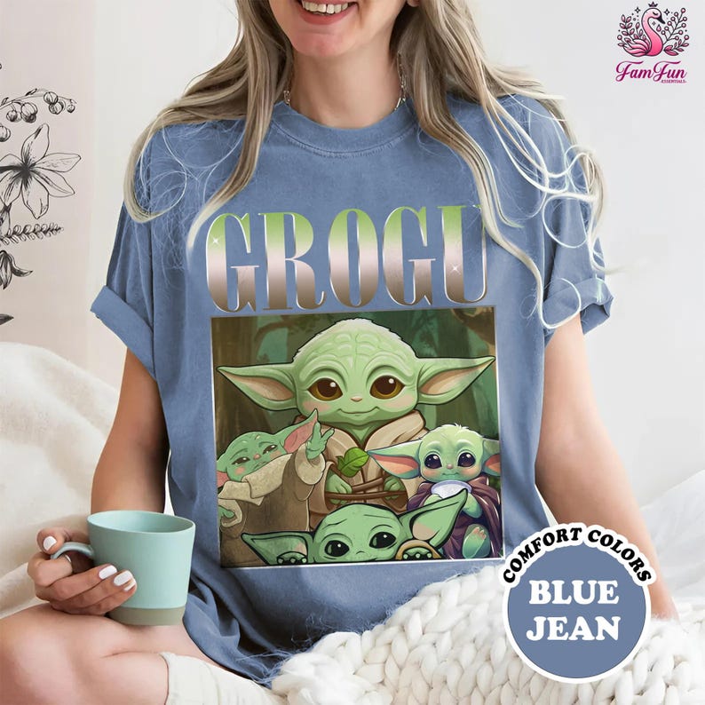 A woman wearing a blue t-shirt with a graphic of a young alien character, likely representing the character Grogy from the Star Wars franchise. She is sitting on a bed and holding a cup, with a cozy, comfortable setting.