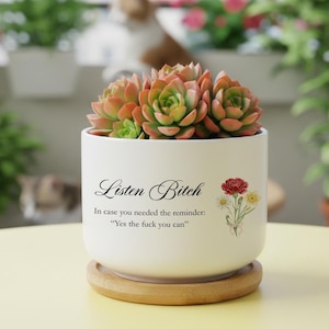Puede incluir: Una maceta de cerámica blanca con una planta suculenta. La maceta tiene el texto "Listen Bitch" y "In case you needed the reminder: 'Yes the fuck you can'". Una ilustración floral está a la derecha. La maceta está sobre un platillo de madera.