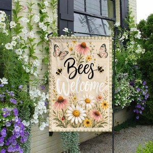May include: A beige garden flag with the words "Bees Welcome" in black and white script. The flag features illustrations of butterflies, bees, and various flowers, including daisies and coneflowers. The flag has a gingham border.