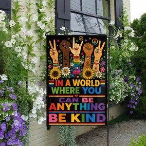 Könnte beinhalten: Eine schwarze Gartenflagge mit dem Text "IN A WORLD WHERE YOU CAN BE ANYTHING BE KIND" in Regenbogenfarben. Die Flagge zeigt Illustrationen von Händen mit verschiedenen Hauttönen, Friedenszeichen und floralen Akzenten.