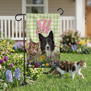 May include: A garden flag featuring a cat and a dog surrounded by colorful flowers. The flag has a green and white checkered background with the letter "Y" in pink, adorned with floral accents. A calico cat walks on the grass.