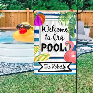Puede incluir: Una bandera de jardín con el texto "Welcome to Our POOL" y "The Roberts" sobre un fondo blanco con rayas azules. La bandera presenta un balón de playa, chanclas y un flamenco rosa. Un gran pato de goma amarillo con gafas de sol flota en una piscina.