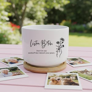 May include: White ceramic planter with the text "Listen B*tch" and "Don't let any motherf*cker disturb your peace." A floral illustration is on the right. The planter sits on a wooden coaster, with photos scattered around.
