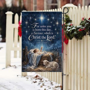 May include: A rectangular garden flag featuring a dark blue night sky with a bright star and the text "For unto you is born this day... a Saviour, which is Christ the Lord." A baby lies in a wooden manger with a lamb resting on the straw below.