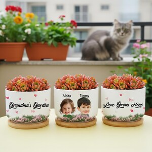 May include: Three white ceramic plant pots with succulents, each on a wooden base. One pot reads "Grandma's Garden", another features photos of two children, and the third says "Love Grows Here". The pots are decorated with hearts.