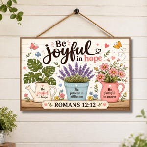 Peut inclure: Une plaque murale rectangulaire suspendue par une corde, avec le texte Be joyful in hope, Be patient in affliction, Be faithful in prayer et ROMANS 12:12. Le motif inclut des illustrations d'un monstera, de lavande et de fleurs roses.
