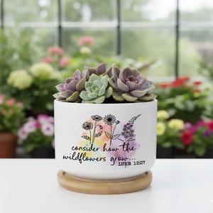 May include: A white ceramic planter with a bamboo base containing a cluster of green and purple succulents. The planter features a colorful watercolor splash design with black line art of wildflowers and the text "consider how the wildflowers grow... LUKE 12:27".