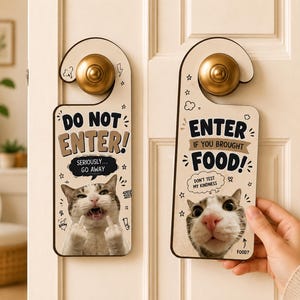 Op de afbeelding: Twee decoratieve deurhangers met kattenafbeeldingen en humoristische tekst. Eén leest "DO NOT ENTER!" met een kat die de middelvinger opsteekt. De andere zegt "ENTER IF YOU BROUGHT FOOD!" met een close-up kattengezicht.