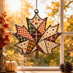 May include: A hanging maple leaf ornament featuring a patchwork design of various autumn-themed fabrics including plaid, gingham, and floral patterns in shades of orange, red, green, and cream, enclosed within a dark metal frame with a chain.