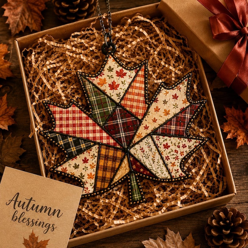 Peut inclure: Un ornement en forme de feuille d'&eacute;rable avec un motif patchwork compos&eacute; de tissus aux couleurs automnales, incluant des carreaux rouges, verts et bruns, le tout cercl&eacute; de m&eacute;tal sombre. Une carte porte l'inscription Autumn blessings.