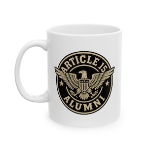 May include: White ceramic mug featuring a black and gold emblem with an eagle, stars, and the words "ARTICLE 15 ALUMNI". The mug has a curved handle and a glossy finish, suitable for hot beverages.