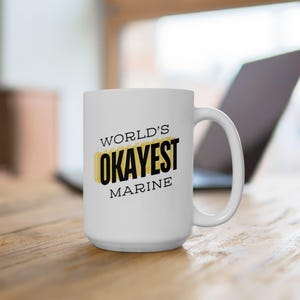 May include: White ceramic mug with the text "WORLD'S OKAYEST MARINE" in black and yellow lettering. The mug has a large handle and is sitting on a wooden surface. A laptop is visible in the background.