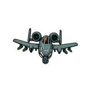 Könnte beinhalten: Ein Aufkleber im Cartoon-Stil eines grauen A-10 Thunderbolt II Flugzeugs. Das Flugzeug hat ein bedrohliches Gesicht mit scharfen Zähnen und einer großen Kanone. Der Aufkleber hat einen weißen Rand und eignet sich für Laptops, Wasserflaschen oder Notizbücher.