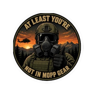 May include: Round sticker featuring a soldier in a gas mask giving a thumbs up. The background shows a sunset, mountains, and a helicopter. Text reads "AT LEAST YOU'RE NOT IN MOPP GEAR."