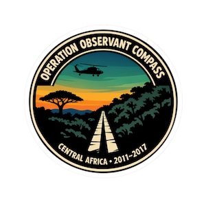 Op de afbeelding: Ronde sticker met een landschapsontwerp. De sticker toont een weg die naar de horizon leidt, een helikopter en de tekst "OPERATION OBSERVANT COMPASS CENTRAL AFRICA 2011-2017". De achtergrond toont een zonsondergang.