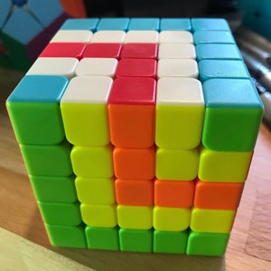 May include: A 4x4x4 Rubik's Cube puzzle with green, yellow, orange, red, white, and blue squares. The cube is in a partially solved state.