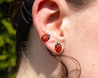 May include: Two small stud earrings shaped like ladybugs. Each earring features a bright red body with distinct black spots, a black head, and small white accents. The ladybug studs are positioned in the earlobe, one above the other, displaying a detailed insect design with a glossy finish and a rounded, compact shape.