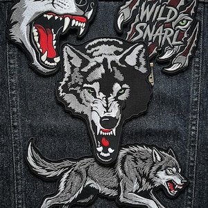 May include: Embroidered patches featuring wolf designs. The patches include a snarling wolf head, a wolf with the text "WILD SNARL", and a running wolf. The patches are black, white, and gray with red and green accents.