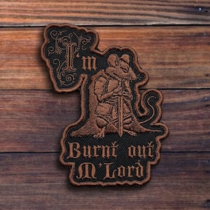 May include: Embroidered patch featuring a brown rat in armor holding a sword, with the text "I'm Burnt out M'Lord" in a gothic font. The patch has a black border and is set against a wooden background.