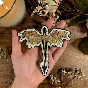 May include: Embroidered dragon patch with gold and green wings, black body, and white border. The patch is held in a hand, showcasing the intricate details of the design. Perfect for adding a unique touch to clothing or accessories.