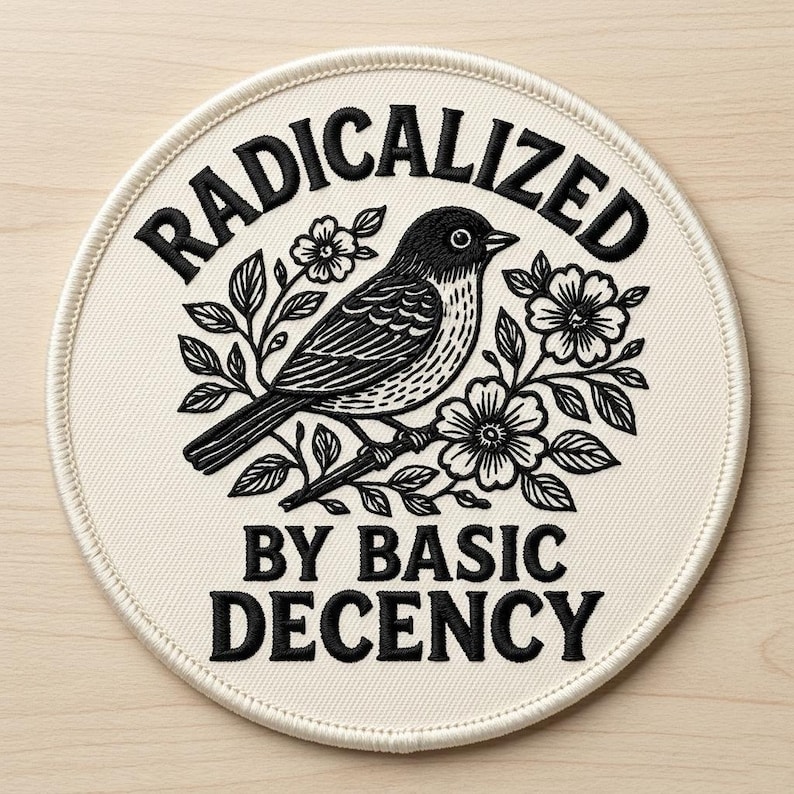 Puede incluir: Parche redondo con el texto "RADICALIZED BY BASIC DECENCY" en negro, rodeando una ilustraci&oacute;n en blanco y negro de un p&aacute;jaro posado en una rama florecida. El parche tiene un fondo color crema y un borde cosido.
