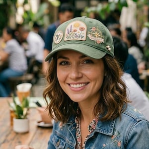 Puede incluir: Gorra de b&eacute;isbol verde con parches bordados que representan un sol, flores y una br&uacute;jula. La gorra tiene una visera curva y est&aacute; adornada con peque&ntilde;os adornos perlados. La palabra "Wanderlust" es visible.