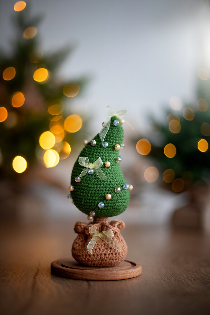 May include: A green crocheted Christmas tree with a brown crocheted base and a yellow ribbon. The tree is decorated with white and blue beads.