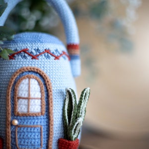 Pu&ograve; includere: Una teiera in crochet blu a forma di casa con una finestra bianca e una porta marrone. La teiera ha una pianta in crochet rosso e bianco in un vaso sul lato.