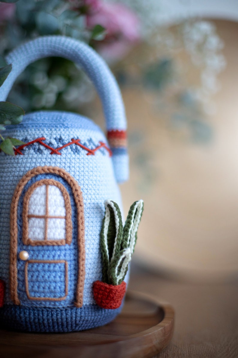 Pu&ograve; includere: Una teiera in crochet blu a forma di casa con una finestra bianca e una porta marrone. La teiera ha una pianta in crochet rosso e bianco in un vaso sul lato.