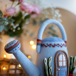 Pu&ograve; includere: Un annaffiatoio in crochet blu a forma di casa con una porta e finestre. L'annaffiatoio ha un manico marrone e un beccuccio. Ci sono due piante in crochet verdi in vasi rossi sul davanti dell'annaffiatoio.