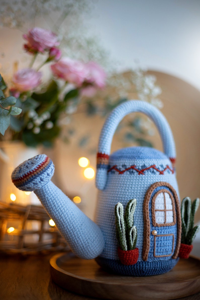 Pu&ograve; includere: Un annaffiatoio in crochet blu a forma di casa con una porta e finestre. L'annaffiatoio ha un manico marrone e un beccuccio. Ci sono due piante in crochet verdi in vasi rossi sul davanti dell'annaffiatoio.