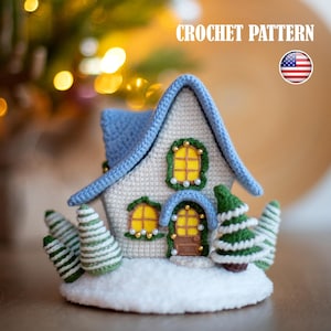 Può includere: Una scena di villaggio invernale all'uncinetto con una casetta. La casa ha un tetto blu e pareti bianche, finestre gialle e una porta marrone. Piccoli alberi all'uncinetto e una base innevata completano la scena. Il testo "CROCHET PATTERN" è visualizzato.