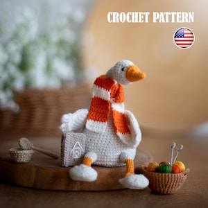 May include: A handmade crochet goose wearing an orange and white striped scarf. The goose has an orange beak and feet. A small crochet house and basket of yarn are next to the goose. The text "CROCHET PATTERN" is at the top.
