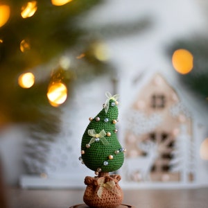 May include: A green crocheted Christmas tree with yellow ribbon bows and pearl beads, standing on a brown burlap sack base with a wooden stand.