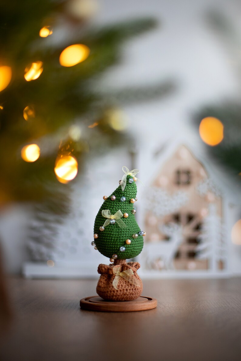 May include: A green crocheted Christmas tree with yellow ribbon bows and pearl beads, standing on a brown burlap sack base with a wooden stand.