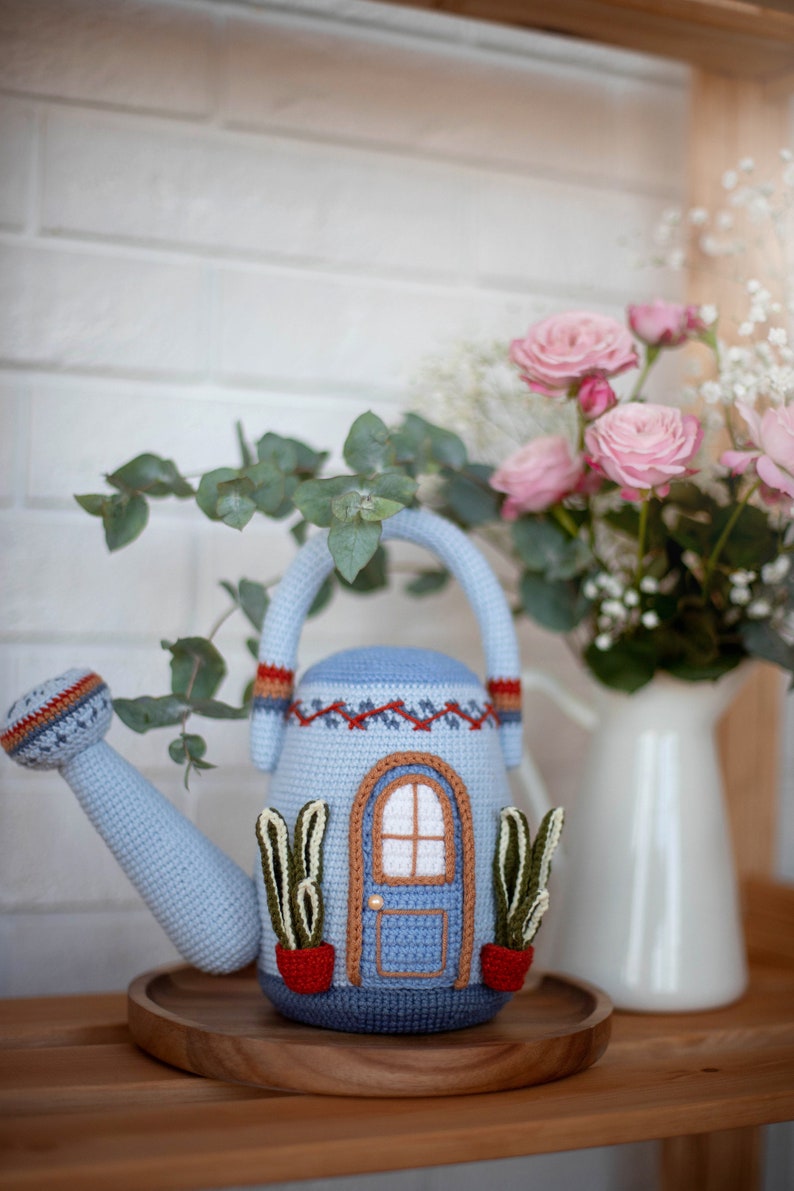 Pu&ograve; includere: Un annaffiatoio di uncinetto blu a forma di casa con una porta blu e due piccole piante in vaso. L'annaffiatoio &egrave; su un vassoio di legno.