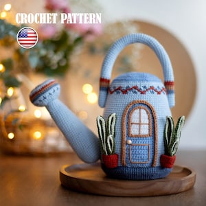 Pu&ograve; includere: Un annaffiatoio in crochet blu a forma di casa con una porta blu e due piccole piante in vaso. L'annaffiatoio ha un manico e un beccuccio marroni.