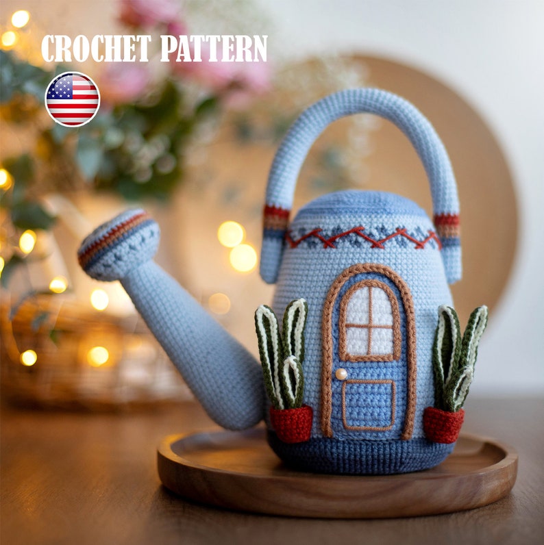Pu&ograve; includere: Un annaffiatoio in crochet blu a forma di casa con una porta blu e due piccole piante in vaso. L'annaffiatoio ha un manico e un beccuccio marroni.