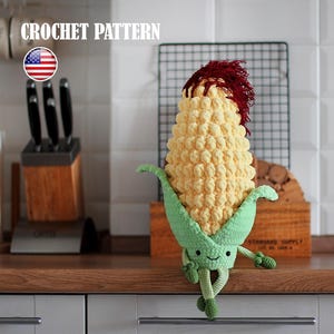 Puede incluir: Un peluche de mazorca de maíz tejido a ganchillo con una cara sonriente, hojas verdes y pelo rojo. El texto "CROCHET PATTERN" está encima del maíz. El peluche está sobre una superficie de madera.