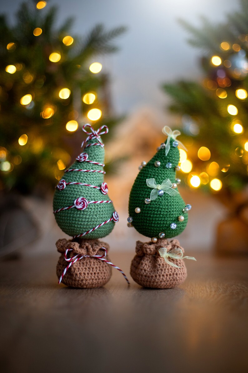 May include: Two crocheted Christmas trees with brown burlap bases. One tree is decorated with red and white twine and the other with white beads and a yellow ribbon.