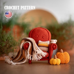 Può includere: Una poltrona all'uncinetto con una coperta e un cuscino, seduta su una superficie di legno con due zucche all'uncinetto. La poltrona è rossa, la coperta è marrone e bianca, il cuscino è beige con accenti gialli, e le zucche sono arancioni e gialle.